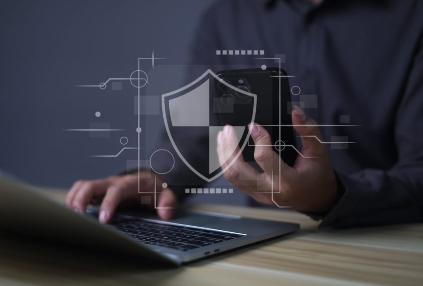 A person holding a smartphone while typing on a laptop with a digital security shield overlay.